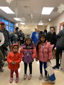 Three young girls stand in the LCH West Grove break room with their new coats as parents and volunteers look on.